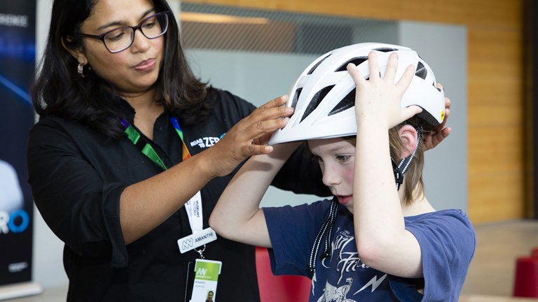 A museum staff member helps a young boy who is trying on a bike helmet.
