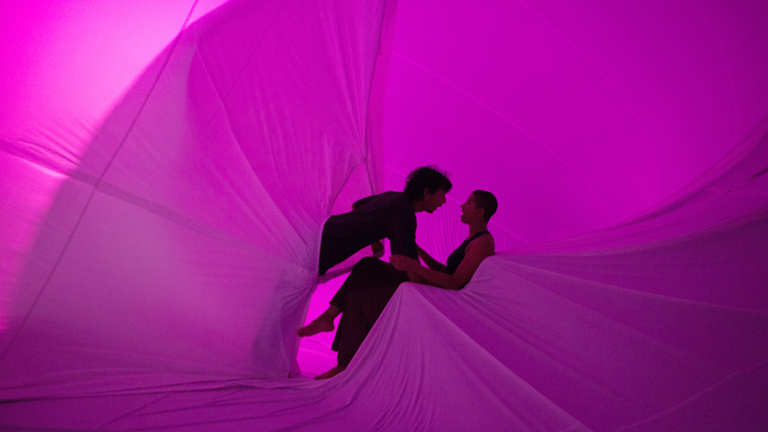 Two dancers interact inside a giant translucent inflatable structure, backlit by pink lighting.