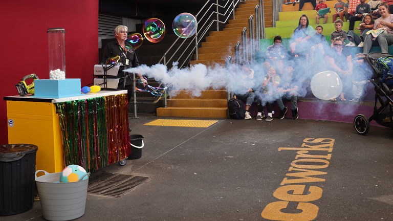 A performer stands behind a fog machine that has made a get of white fog. There are a few bubbles floating around. One has been filled with the white fog. A crowd of people, adults and children, are sitting in a tiered amphitheatre and watching on. The tiered seating is painted in rainbow colours.
