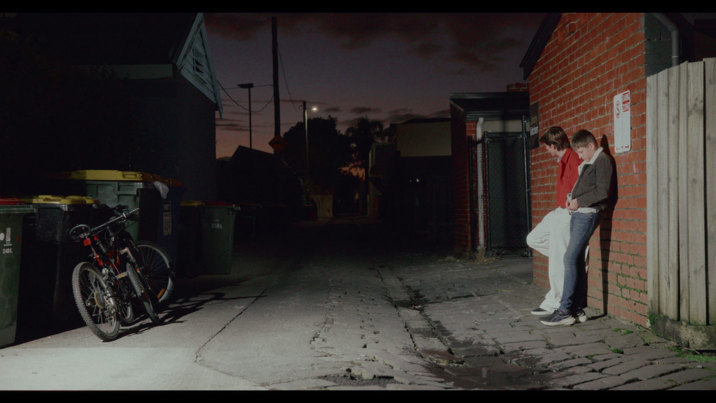 Film still showing two teenaged boys standing in a laneway at nightfall, leaning against a brick building, with a pair of bikes propped up against nearby garbage bins.