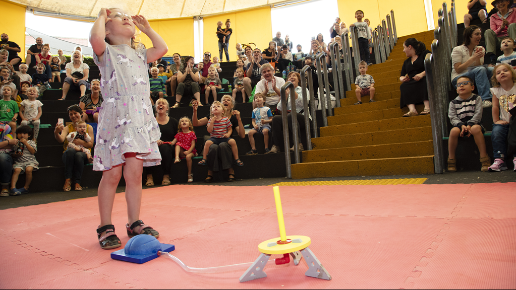  A child stands next to a stomp rocket, looking upwards with safety goggles on. A crowd behind the child looks on in excitement.