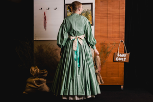 Woman in long green dress with a white apron, sitting in front of a wooden backdrop with a framed painting, hanging animal carcass, dried plants, and a "Staff Only" sign. 