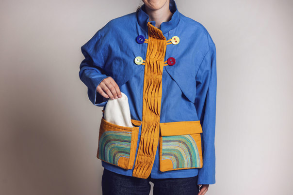 Model wearing a blue jacket with colourful oversized buttons and an orange pleated placket, featuring pockets with decorative embroidery.