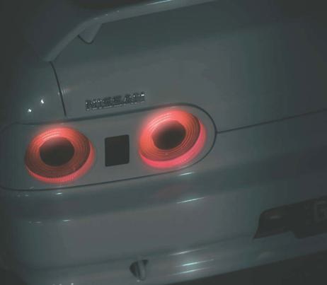 Close-up of a light-coloured Nissan R32 Skyline car's rear panel, showing two illuminated, circular red tail lights, photographed in low lighting.