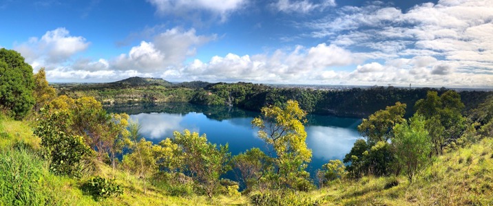 A lake surrounded by bush, with a mountain range in the background. There are clouds in a blue sky.