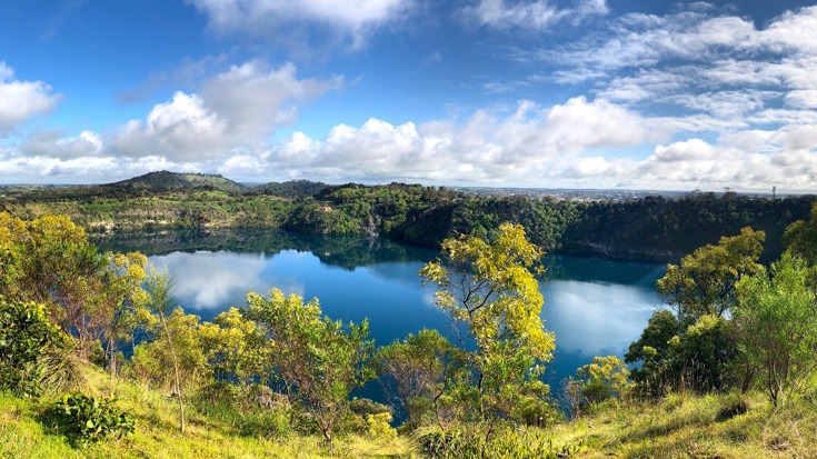A lake surrounded by bush, with a mountain range in the background. There are clouds in a blue sky.