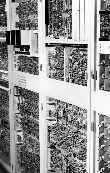 Black and white photo of a man in a suit operating a very old computer which is several cabinets in a row with many wires, switches and dials. 