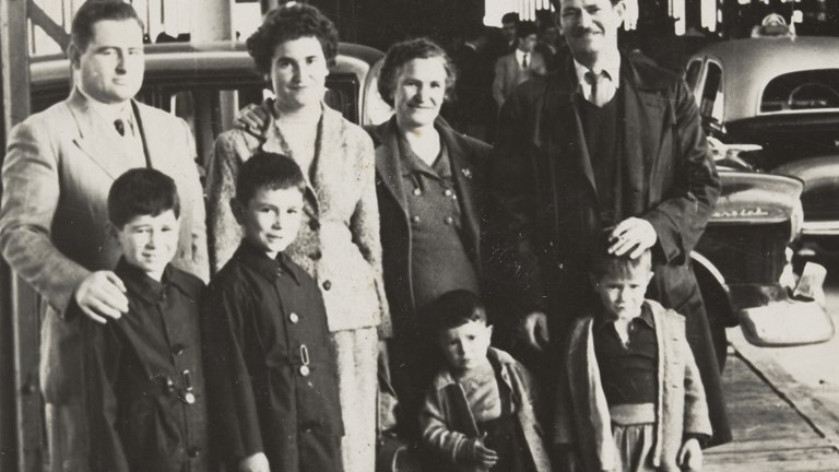 Extended family standing around suitcase on Station Pier, under a large roofed structure. Two men, two women, two boys and two infant children. There is a large suitcase in the foreground with rugs strapped to the side. Cars are parked in the background.