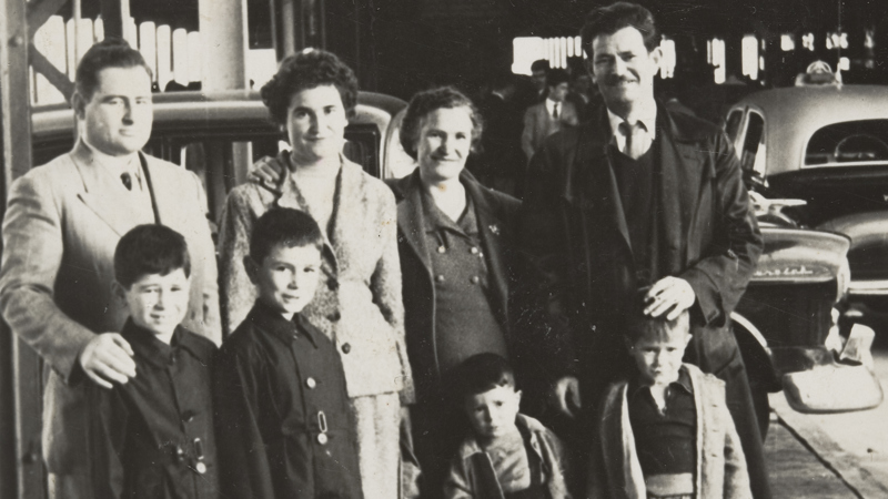 Extended family standing around suitcase on Station Pier, under a large roofed structure. Two men, two women, two boys and two infant children. There is a large suitcase in the foreground with rugs strapped to the side. Cars are parked in the background.