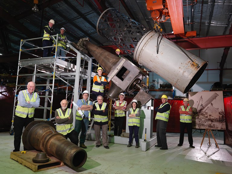 A group of people wearing hi-vis vests in engineering workshop posing with parts of a  historic telescope