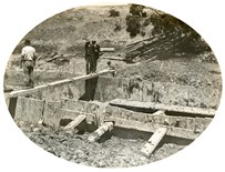 Three men standing on a beam across the coffer dam of Pier 3 of the permanent bridge over Woady Yallock Creek on the Gheringhap to Maroona line, 1912