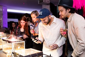 People holding drinks peering in display boxes. One person is taking a photo with a phone