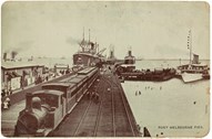 A passenger train at Port Melbourne Pier. Some ships moored at pier. People walking along the side of the pier. A number of people are wading in the shallow water. A ship on the right is displaying bunting.