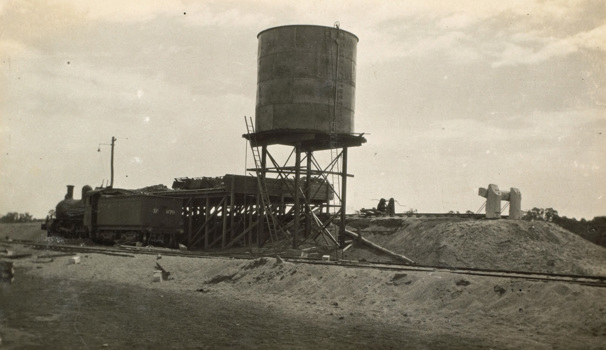 A steam locomotive beside a coal and tank stage at Balranald, circa 1927. There are railway tracks at a higher level behind the 24,000 gallon (109.2kl) water tank. Balranald was the last station of the Moama to Balranald line.