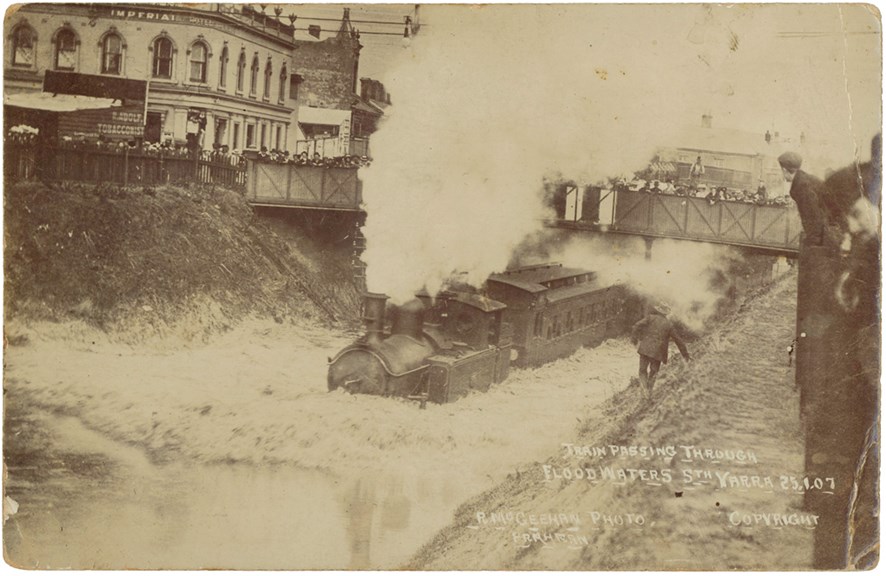 Steam engine with passenger carriages attached passing through a flooded railway cutting in South Yarra, 25 January 1907. The Imperial Hotel is in the background.