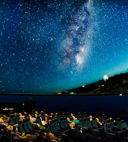 People sitting in theatre recliner chairs looking at a projection of the night sky