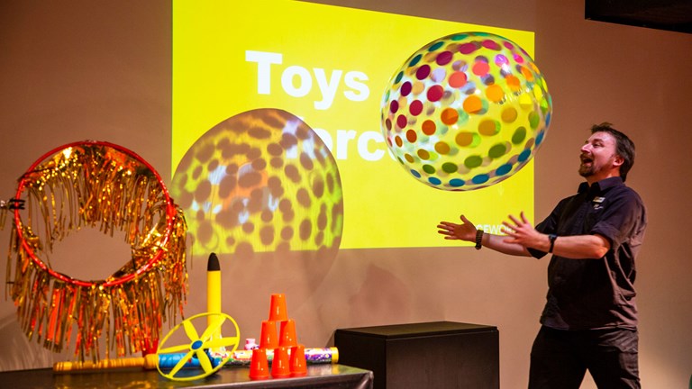A presenter throws a giant beach ball in the air. There are other toys on the table including a foam rocket, a hula hoop with glittery strands, a stack of red plastic cups and a helicopter.  