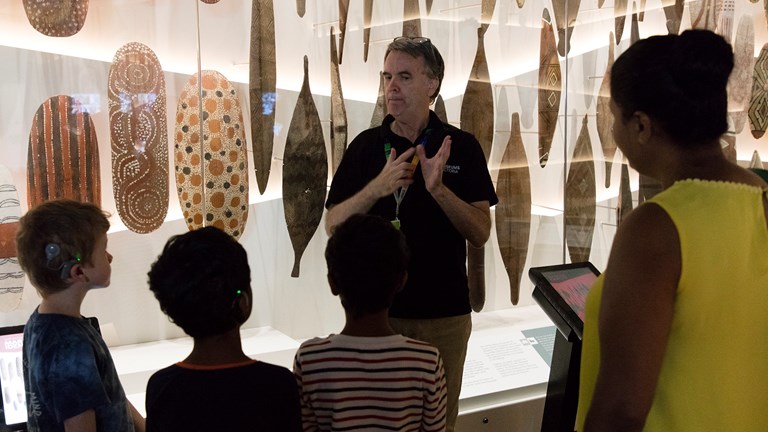 Man doing a presentation  in Auslan to a small group of people in front of a showcase displaying Aboriginal sheilds
