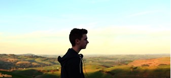 Film still showing the side profile of a young man, with headphones around his neck, standing above an expanse of rolling green hills in the evening sun