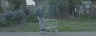 Film still shows the side profile of a girl riding on the back of a shopping trolley down the street outside suburban homes