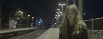 Film still shows a young girl with long brown hair stands alone, looking troubled, at a train station at night time