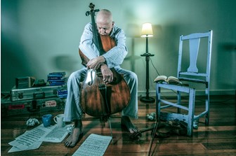 A seated older man is dressed in his pyjamas while sadly holding a cello, with his chin resting on the top of the instrument, looking contemplatively at his sheet music on the ground. Books, music, and mugs are strewn around him. The photograph comprises mainly blue/grey objects, except for the cello, and an overall blue colour pallete