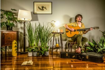 A young man is seated, surrounded by plants, looking up pensively as he plays his guitar.  The photograph comprises mainly yellow/green objects objects and has an overall yellow colour pallete