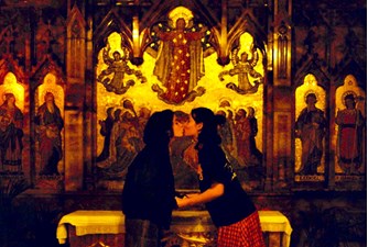 Two alternatively-dressed young girls kiss holding hands, kissing on the lips, at the centre of the golden altar in a church.  Their bodies and the ornate church background create symmetrical shapes.
