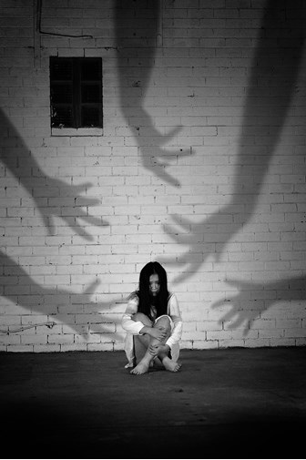 A black and white photograph of a fearful girl with long black hair, wearing a short white dress, sitting cross-legged on the ground, holding her knees to her chest.  She is an empty room with white brick walls.  Large shadows of outstretched arms are reaching towards her, as if trying to grab her