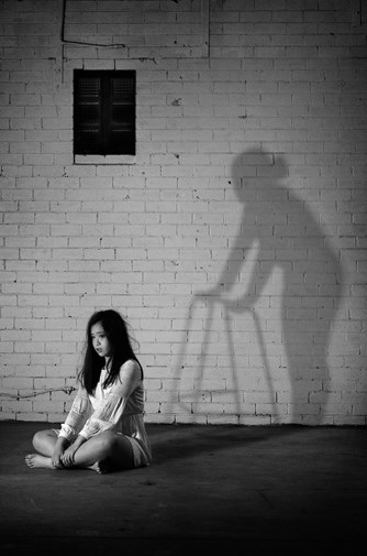 A black and white photograph of a fearful girl with long black hair, wearing a short white dress, sitting cross-legged on the ground.  She has her back turned away from the shadow of an elderly figure with a zimmer-frame 