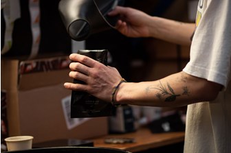 A close up of a tattooed arm puring ground coffee into black packaging