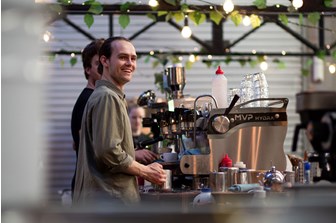 A young man making coffee at a coffee machine smiles in the direction of the photographer, seemingly unaware of the image being taken