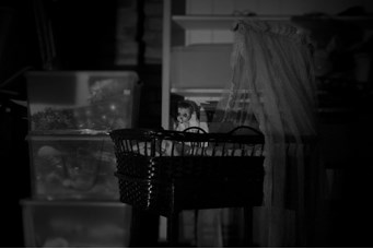 A black and white photo of an old-fashioned wicker bassinet with a lace canopy. An old-fashioned doll sits upright, with her arm upwards. 
