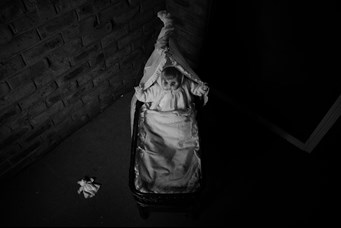 A black and white aerial photo of a an old-fashioned doll in a wicker bassinet, with both arms positioned upwards. A discarded toy lies to the left. 