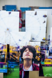 A collage of mulitple hanging photographs that show a young boy in a red hoodie looking upwards. In the background is an empty fairground with a yellow ferris wheel and blue tables. 