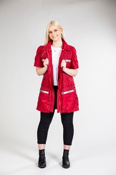 A blonde girl wears a red jacket. The arms have been zipped off to create short sleeves. A line of reflective material is used across the top of the four front pockets