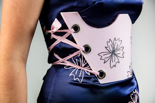 A close up of a pink resin brace that is secured using pink ribbon over a blue satin dress with pink floral detailing.