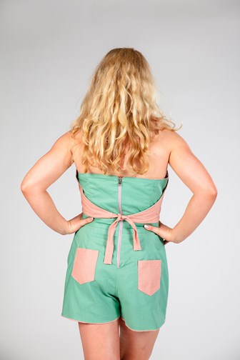 A model is seen from the back wearing a short green playsuit. There are two pink pockets on the shorts and a pink tie is seen from the back