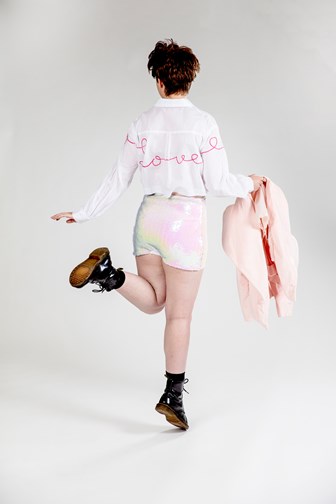 A girl is seen dancing from behind. She is wearing sequined shorts, a cotton long-sleeved shirt with the woerd 'love' embroidered on the back. She is holding a pink velvet jacket. 