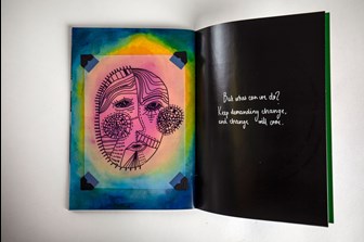 A photograph of an open art zine showing two pages, one is a colourful page with a hand-drawn illustration of a face on transparent acetate, the other is a black page with white text, in a hand-written style