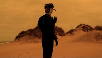 A man wearing black protective gear, including gloves and a gas mask, holds up a black box while standing on an empty beach. The light is dim, yellow and hazy