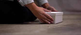 A close up of a pair of hands placed either side of a white box that is sitting on a concrete floor. The person is kneeling and their sleeves are rolled up to reveal a white shirt. 