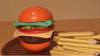 A burger made from everyday objects, such as bottle caps for pickles and green latex gloves for lettuce leaves. Thick yellow pencils are used to represent fries, which are placed to the right of the burger. 