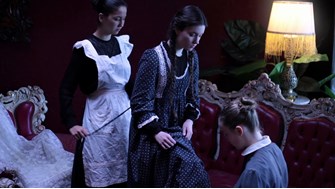 A girl with long dark pigtails stands between two maids who are dresssing her in a dark room lit only by a lamp. A lace dress lies on an ornate red chair in the background. 