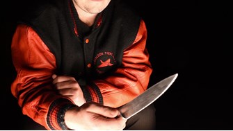 A close up of a young man in a navy and red bomber jacket holds a large kitchen knife in the dark., his face is out of frame