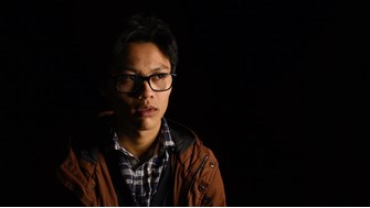 A teenage boy wearing glasses, a check shirt and a brown coat stands alone in the dark, with an apprehensive expression 