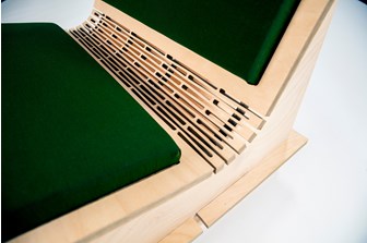 A close up of the curve in between the top and bottom of a plywood chair, showing detailed laser-cut lines and a green cushion