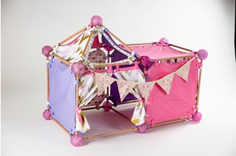 Overall view of a children's play area.  It is modular in it's form and is constructed with wood poles and resin balls, with pink, purple and patterned fabric panelling.  Bunting with childrens drawings on hangs across the front.