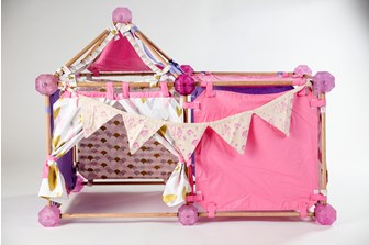 Front view of a children's play area.  It is modular in it's form and is constructed with wood poles and resin balls, with pink, purple and patterned fabric panelling.  Bunting with childrens drawings on hangs across the front.