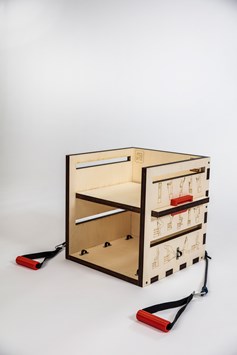 An overall shot of the 'Gym Box', an exercising tool made from wood.  It is a wooden cube with lasercut drawings of people doing different exercises on the sides. Black and red resistance bands are fixed to either side.